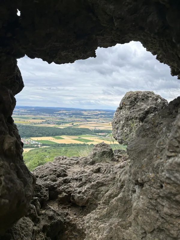 Blick durch eine Felsöffnung am Staffelberg auf eine weite, bewölkte Landschaft mit Feldern und Ortschaften. Im Vordergrund rahmen grobe Felsformationen die Aussicht ein. (KI-generierte Bildbeschreibung)