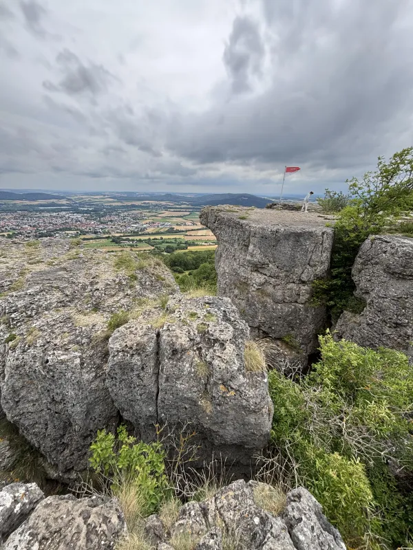Blick von der markanten Felsformation am Staffelberg auf eine weitläufige Landschaft mit Feldern und Ortschaften. Auf dem Felsvorsprung steht eine Person neben einer kleinen rot-weißen Flagge unter einem bewölkten Himmel. (KI-generierte Bildbeschreibung)