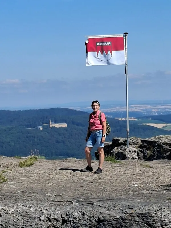 Eine Wanderin steht auf dem felsigen Plateau des Staffelbergs neben einer wehenden Frankenflagge. Im Hintergrund ist bei klarem Wetter die Basilika Vierzehnheiligen in der hügeligen Landschaft zu sehen. (KI-generierte Bildbeschreibung)