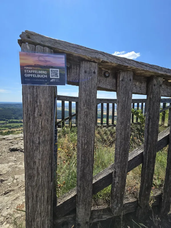 An einem hölzernen Aussichtsgeländer am Staffelberg ist ein Hinweisschild mit einem QR-Code zum digitalen Gipfelbuch befestigt. Im Hintergrund ist bei blauem Himmel die weitläufige Landschaft zu sehen. (KI-generierte Bildbeschreibung)