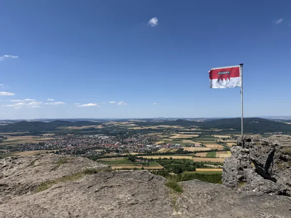 Blick vom felsigen Plateau des Staffelbergs über eine weitläufige Landschaft mit Feldern und Ortschaften. Auf dem Fels steht eine Flagge mit dem fränkischen Rechen vor einem blauen Himmel. (KI-generierte Bildbeschreibung)