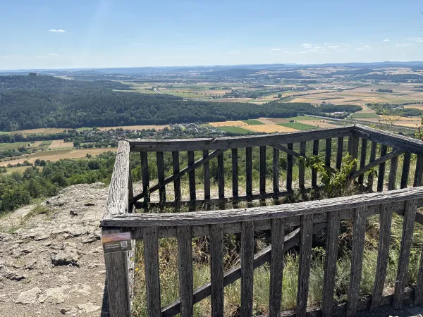 Blick von einer hölzernen Aussichtsplattform auf dem Staffelberg über ein weitläufiges Tal mit Feldern, Wäldern und Ortschaften. An einem der Pfosten ist ein Schild mit dem Hinweis auf ein digitales Gipfelbuch angebracht. (KI-generierte Bildbeschreibung)