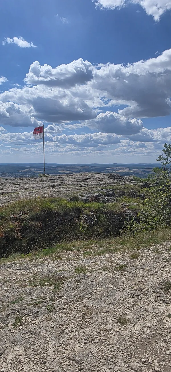 Eine rot-weiße Fahne auf dem felsigen Plateau des Staffelbergs vor weitem Blick über eine hügelige Landschaft bei blauem Himmel mit weißen Wolken. (KI-generierte Bildbeschreibung)