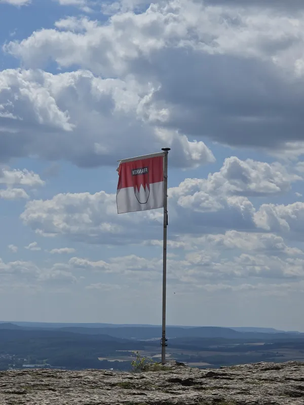 Eine rot-weiße Frankenflagge weht an einem Fahnenmast auf dem felsigen Gipfelplateau des Staffelbergs. Im Hintergrund erstreckt sich eine weite Landschaft unter einem bewölkten Himmel. (KI-generierte Bildbeschreibung)